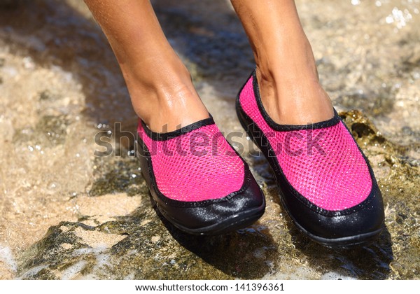 Water shoes / swim shoe in Pink neoprene on rocks\
in water on beach. Closeup detail of the feet of a woman wearing\
bright pink neoprene water shoes standing on rocks at the edge of\
the ocean.