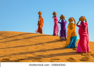 Water Shortage And Crisis Concept, Indian  Women Carrying Heavy Jugs Of Water On Their Head And Walking On Sand Dune In The Hot Summer Desert Against Blue Sky. Environmental Change