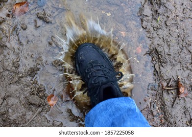 Water Splash From Shoes. Men's Feet In Hiking Shoes Steps Into A Puddle. Rain Footwear For Man Or Woman. Trekker Boots For For Cold And Weather Hike. Pair Of Waterproof Travel Shoe In Blue Jeans.