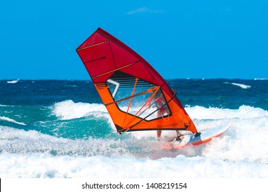 water sports: windsurfer with red and orange sail riding the waves during a sunny summer afternoon.
