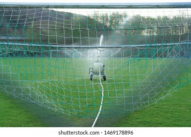 Water spray system on a green football field, seen through goal net.