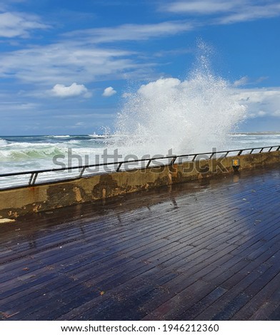 Water sprayed by ricochets splashed off of a big wave breaking against the boardwalk's wall and reflected on the wet wooden deck. Yellow sign signaling caution slippery when wet. Sky. Feather clouds.