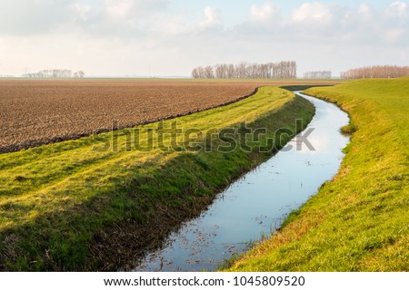 Water surface of a meandering ditch reflects the white clouds in the blue sky. The field next to the ditch has recently been plowed in preparation for the new growing season.