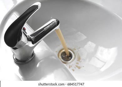 Water tap faucet with flowing contaminated muddy and dirty water in a white bathroom sink.