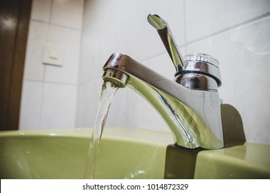 Water tap or faucet with water flowing out of the outlet, mounted on a green sink or wash basin. Old looking bathroom with open tap with clean water.