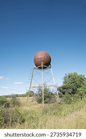 A water tower with a rusty surface rises on metal supports in a grassy field. Surrounding plants and trees grow around the base. The sky shows a few clouds.