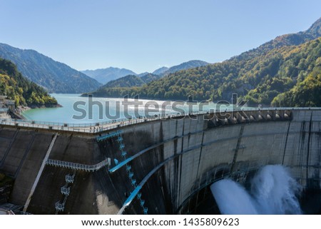 Water Turbines Are Producing Electricity At Power Plant. Panorama View Of Hydro Power Station And People On The Kurobe Lake Dam,Toyama. River Dam and Tateyama Mountains.