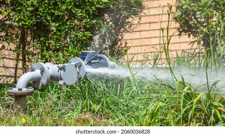 Water Valve Meter High Pressure Broken Leaking Leaking Gasket On Bolted Flanges Spraying Onto Grass Roadside Verge Outdoors.