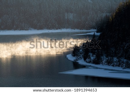 Water vapor in the evening light of sunset on the ice-cold Lake Plansee in winter with a wooded shore