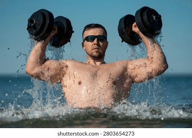 Water workout creates refreshing summer fitness. Man lifting dumbbells in ocean water. Splashing waves show active lifestyle. Athletic body shows vitality and strength.