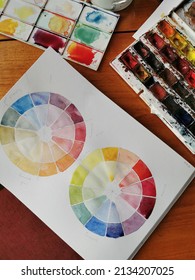 A watercolor painting of two color wheels with primary and intermediary colors, a part of a watercolor pan against a warm toned wooden table - Shutterstock ID 2134207025
