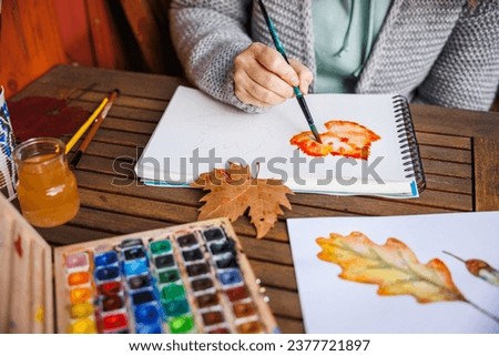 Watercolor painting. Woman artist paints an autumn theme on paper. Palette of colors on the table