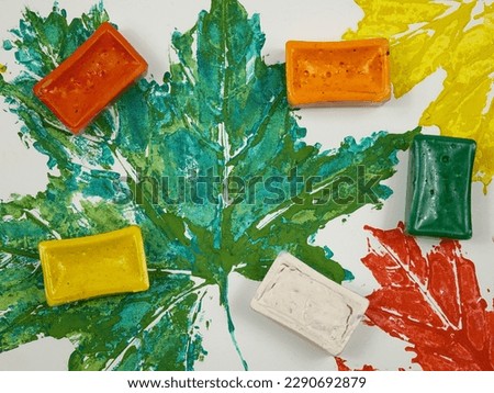 Watercolor paints in plastic containers lie on the drawing of maple leaves in red-yellow autumn tints on the white background  