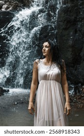 A waterfall is in the background and water flows down the rocks. The woman looks away and has a calm expression on her face.