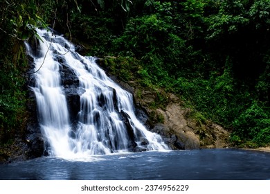 A waterfall cascades down a rocky cliff into a clear pool of water. The waterfall is surrounded by lush green trees, and the sound of the water is calming. - Powered by Shutterstock