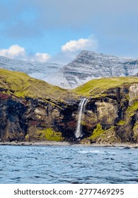 Waterfall cascading down dramatic cliffs into Atlantic Ocean with snow capped mountain, Faroe Islands