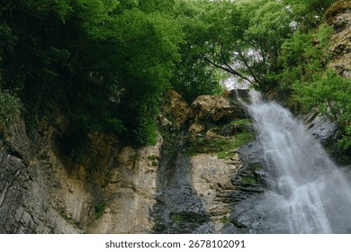 Waterfall cascading down rocky cliffs surrounded by lush green forest in a serene natural landscape with sunlight filtering through dense foliage.