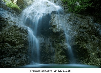 A waterfall cascading into a scenic pool near large rocks and lush green foliage.