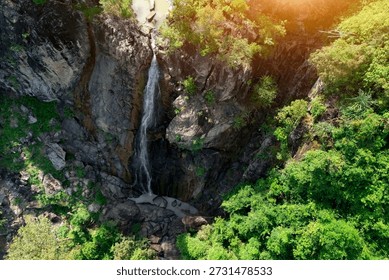 Waterfall drops vertically down a dark, rocky cliff into a small pool. Lush, green vegetation covers the area around the water feature during the daytime.