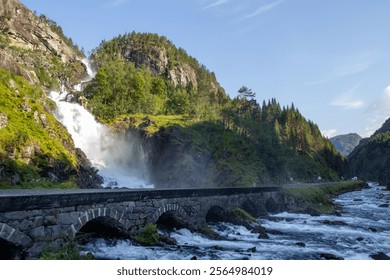 Waterfall flows dramatically over rock formations into a river below, surrounded by vibrant trees and hills. A stone bridge spans the river, enhancing the tranquil landscape.