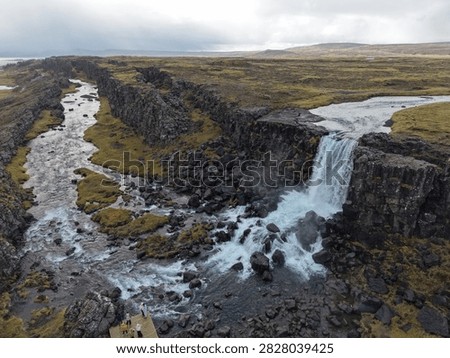 A waterfall flows over black volcanic rocks into a river, surrounded by green moss and cloudy skies.