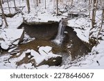 A waterfall flows over an overhang into the valley below. Blue hen Falls is located in Cuyahoga Valley National Park. It is a cold day in winter, and snow blankets the surrounding forest.