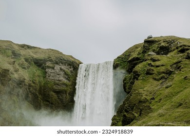 Skógafoss Waterfall in Iceland. Beautiful waterfall and green hills around it on an overcast day. Hiking to a waterfall on a cloudy day in Iceland.