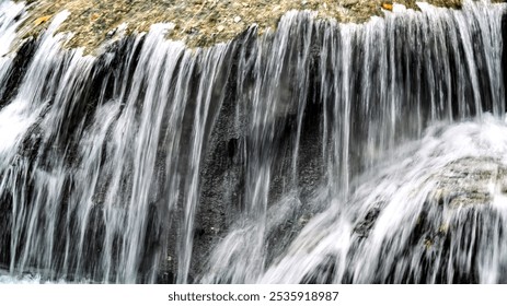 A waterfall is shown in a photo, with the water flowing down a rocky surface. Concept of power and natural beauty, as the water cascades down the rocks. The scene is likely taken in a remote location - Powered by Shutterstock