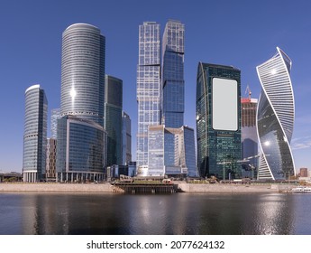 Waterfront cityscape with blank billboard for advertising on the side of a modern glass skyscraper with helical building alongside and continuing development with cranes behind - Shutterstock ID 2077624132