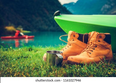 Waterfront Kayak Camping Concept Photo. Kayak And Trail Shoes On The Shore And The Kayaker On The Glacial Lake Water In The Background. 