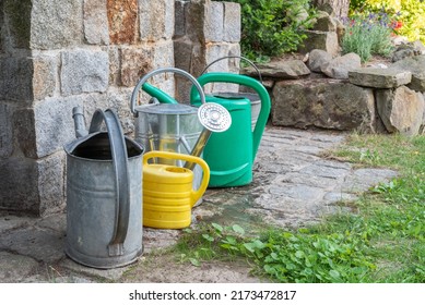 Watering Cans Full Of Water In Front Of Stone Well In Garden During Drought In Summer