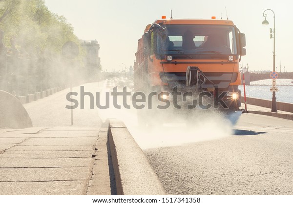 Watering machine washes city road with water. Big\
orange watering machine washes the road part with water. Sun day in\
Saint-Petersburg,\
Russia