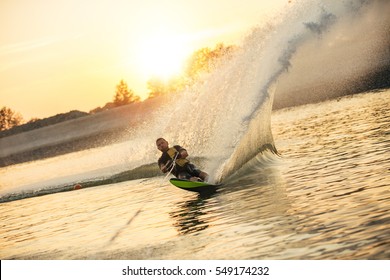 Waterskier moving fast in splashes of water at sunset. Man wakeboarding on a lake