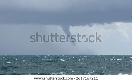 A waterspout, a vortex of cloud touching the oceans surface, is visible under a dark, stormy sky. The sea is choppy with visible waves.