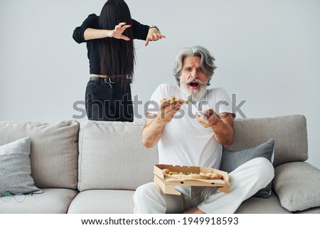 Wathing TV with scary woman with black hair near him. Senior stylish modern man with grey hair and beard indoors.