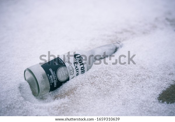 Waukegan, Illinois /\
United States - February 15 2020: Corona beer bottle lying in sandy\
snow covered beach