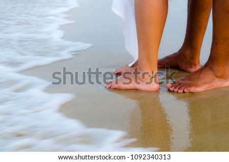 Wave of the sea in the sand reaching the feet of a couple. The wave is foamed. On one foot there is an anchor tattoo.