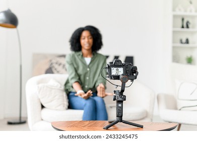 With a wave and a smile, an African-American woman engages her online audience, creating an inviting atmosphere in her vlog from a minimalist living space - Powered by Shutterstock