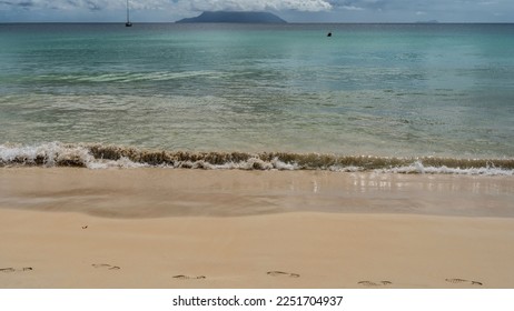 A wave of turquoise ocean foams on the shore, mixing with sand.  Footprints in wet sand. Outlines of the island and yachts on the horizon. Clouds in the blue sky. Seychelles. Mahe. Beau Vallon. - Shutterstock ID 2251704937