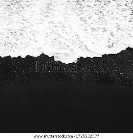 A wave of white foam rolls from above onto a black sandy beach in Bali. Close up aerial view of the sea and top view