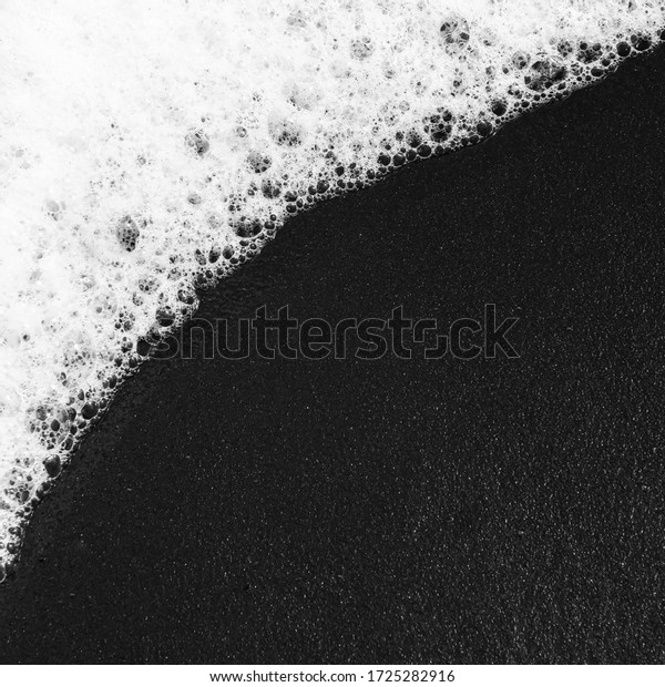 A wave\
of white foam rolls diagonally across the black sandy beach in\
Bali. Close up aerial view of the sea and top\
view