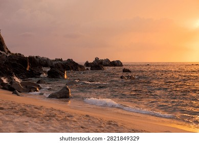 Waves breaking on a sandy beach and rocks at golden sunset, Calabria, Italy. Warm light on the shoreline, sea foam and a rugged rocky headland under a soft orange sky in summer