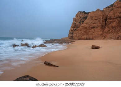 Waves crash against rocks along a deserted beach under a cloudy sky, framed by steep, rugged cliffs - Powered by Shutterstock