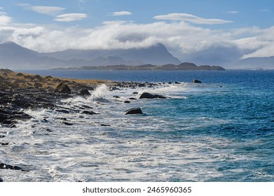 Waves crash against the rocky shore as clouds hover above distant mountains. The serene coastline contrasts with the power of the ocean under a bright blue sky. - Powered by Shutterstock