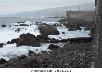 Waves crash against the rocky volcanic shore of Tenerife, with a historic stone wall and distant mountains adding to the dramatic coastal landscape - Powered by Shutterstock