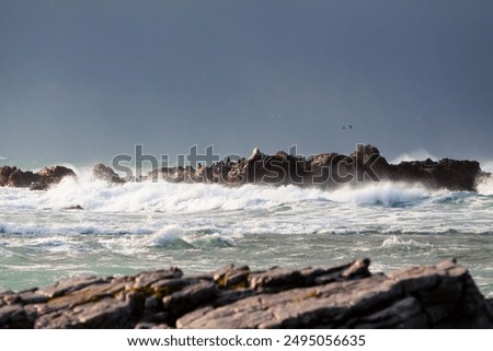 Waves crash dramatically against jagged rocks in the ocean, with mist and spray rising into the air, while the foreground features weathered, rocky terrain and the background is dark and stormy.