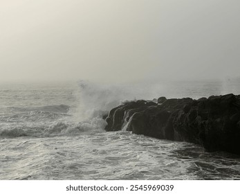 Waves crash dramatically against rocky formations on the rugged west coast under a foggy sky. The powerful water motion and misty atmosphere create a wild, untamed coastal scene that highlights nature - Powered by Shutterstock