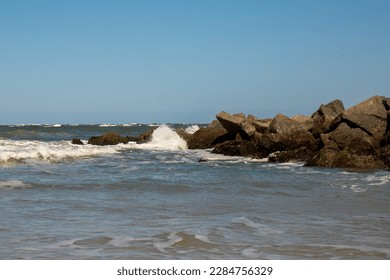 Waves crash on dark rocks with a uniform blue sky above.  White Sea foam and ocean spray and small waves in the foreground.  Waves crash during high tide in Florida on the Atlantic Ocean coastline. - Shutterstock ID 2284756329