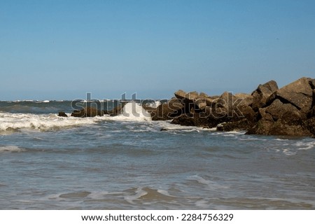 Waves crash on dark rocks with a uniform blue sky above.  White Sea foam and ocean spray and small waves in the foreground.  Waves crash during high tide in Florida on the Atlantic Ocean coastline.