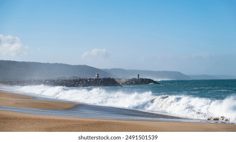 Waves crash onto a sandy beach near a rocky breakwater with red and white lighthouses, under a clear blue sky.  - Powered by Shutterstock